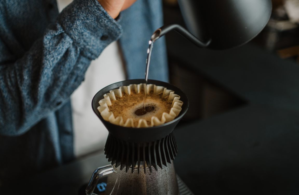 Pouring hot water from a gooseneck kettle into a paper filter cone placed over coffee in a glass carafe, with a person in a blue sweater partially visible in a dimly lit setting.